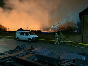 Firefighters carrying hose, fire engine parked on road with smoke in the background