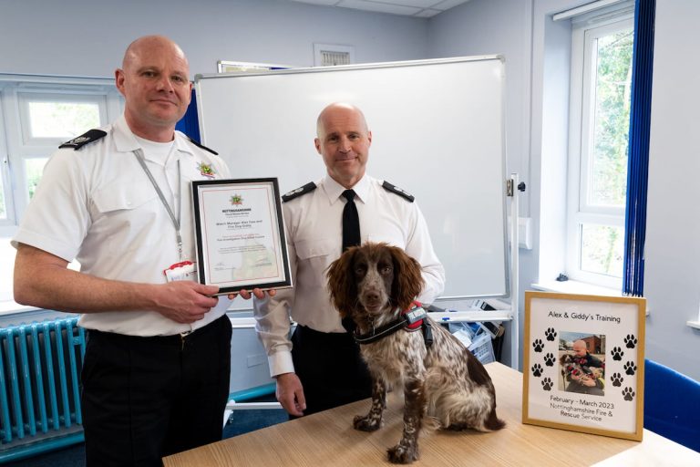 An officer presenting a certificate, with Alex Daw stood next to him and Giddy, the fire dog, sat on a table in front of them