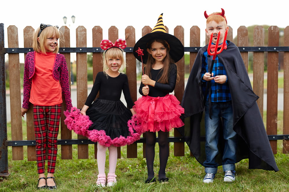 Four children dressed in Halloween costumes in front of a fence.