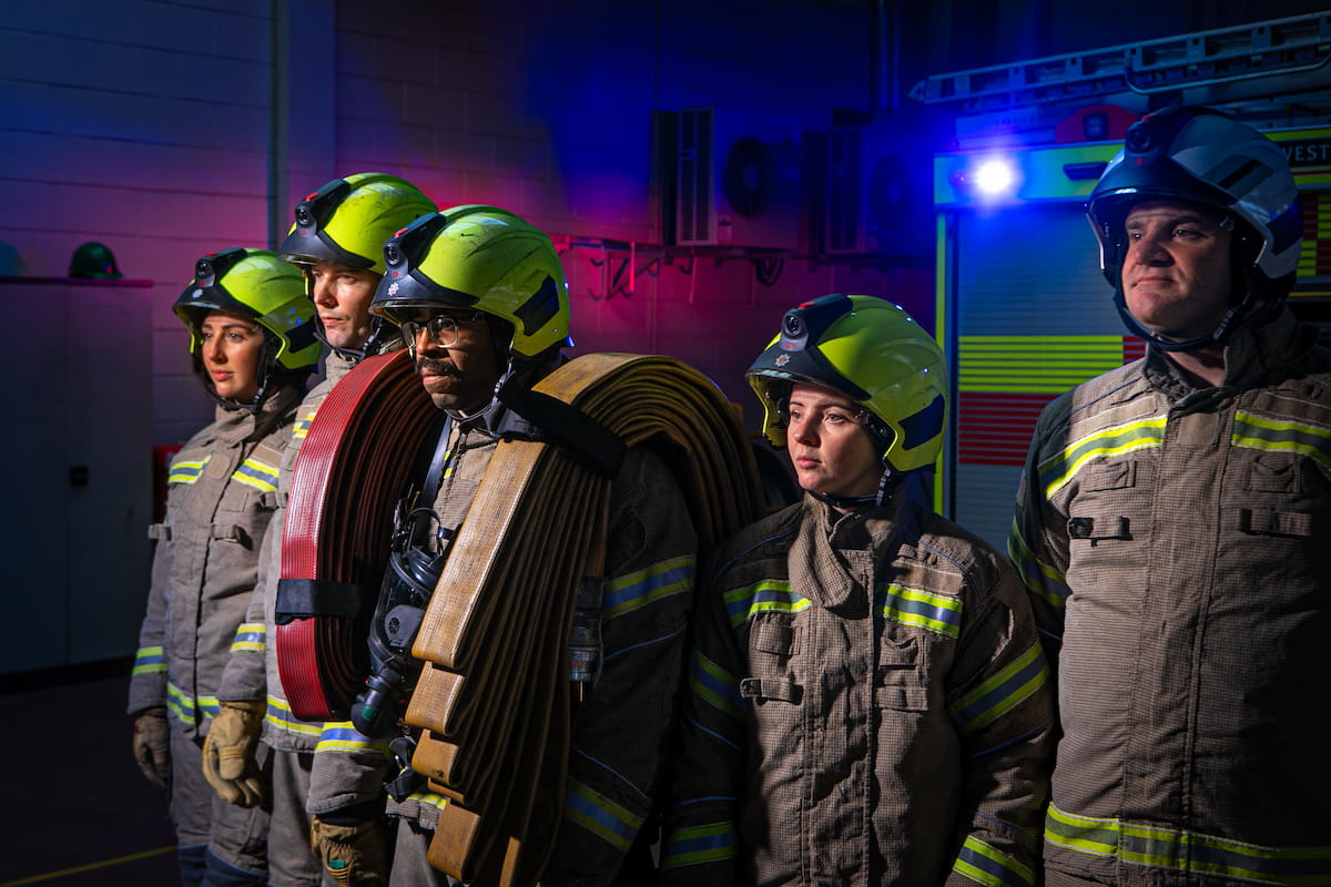 A side view of five firefighters in PPE, with the firefighter in the centre carrying lots of equipment