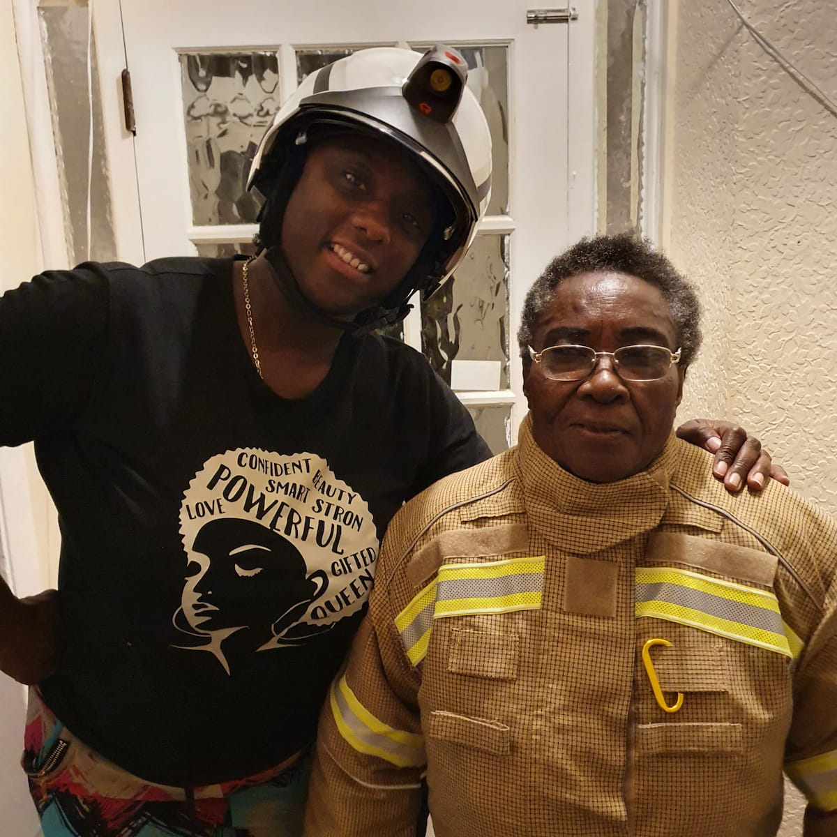 Sam Samuels, dressed in a firefighter’s helmet and casual t-shirt, smiling next to an older woman in firefighter jacket.