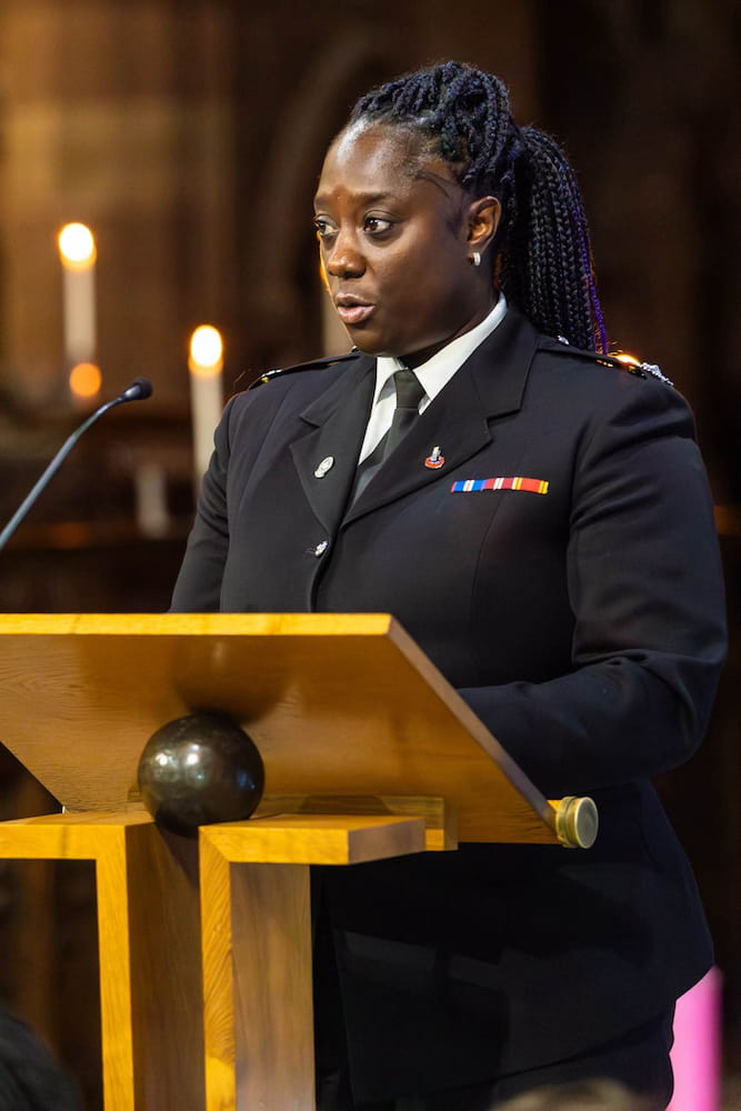 Sam Samuels at a podium in formal fire service dress, speaking at an event with candles glowing softly in the background.