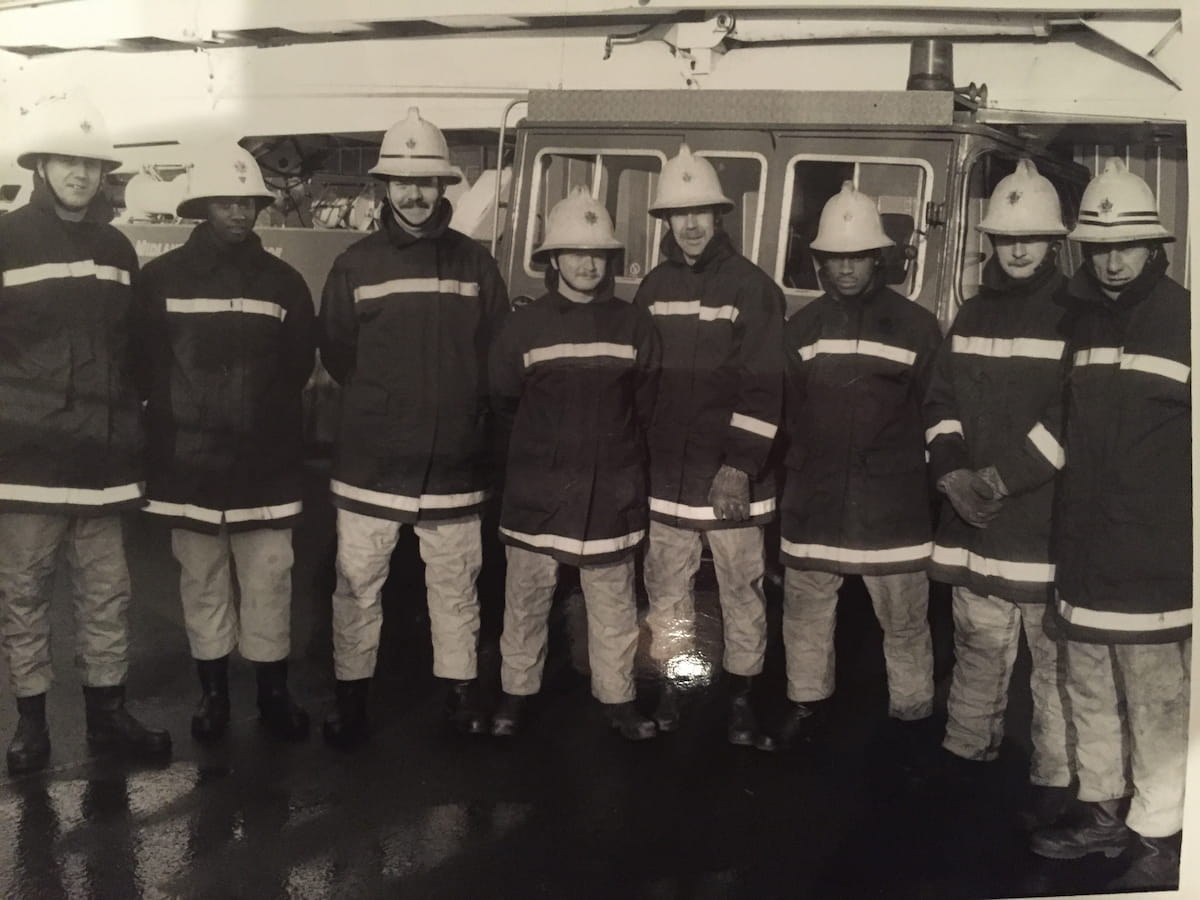 A black-and-white photo of Sam Samuels with seven fellow firefighters, all wearing traditional fire service uniforms, standing together in front of a fire engine, showcasing an early stage in her firefighting career.