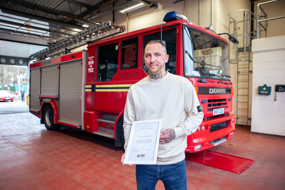 Tom Alexander poses infront of a fire engine holding a framed certificate