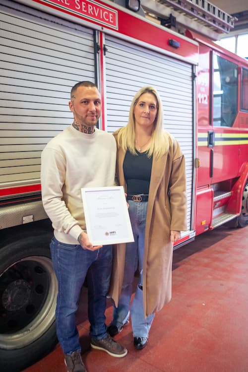 Tom Alexander poses with his partner in front of a fire engine after receiving his Certificate of Meritorious Action at Haden CrossFire Station