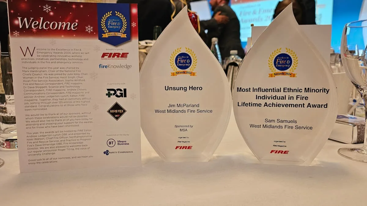 Two award plaques from the Excellence in Fire and Emergency Awards 2024. The left plaque reads "Unsung Hero, Jim McParland, West Midlands Fire Service," sponsored by MSA. The right plaque reads "Most Influential Ethnic Minority Individual in Fire Lifetime Achievement Award, Sam Samuels, West Midlands Fire Service." Both awards are displayed on a table alongside event menus and glasses.