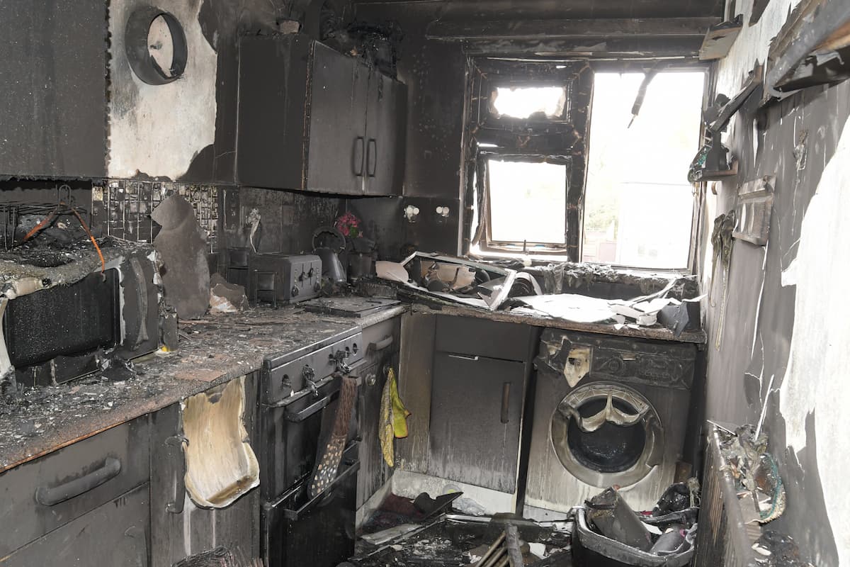 The burnt out remains of the kitchen, with cupboards and washing machine visible, but everything covered in soot, fire and smoke damage